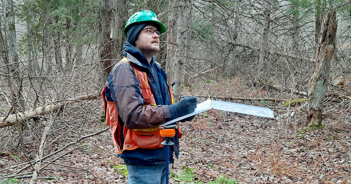 Man in safety vest and green hard hat writing in a large notebook in the middle of a forested landscape